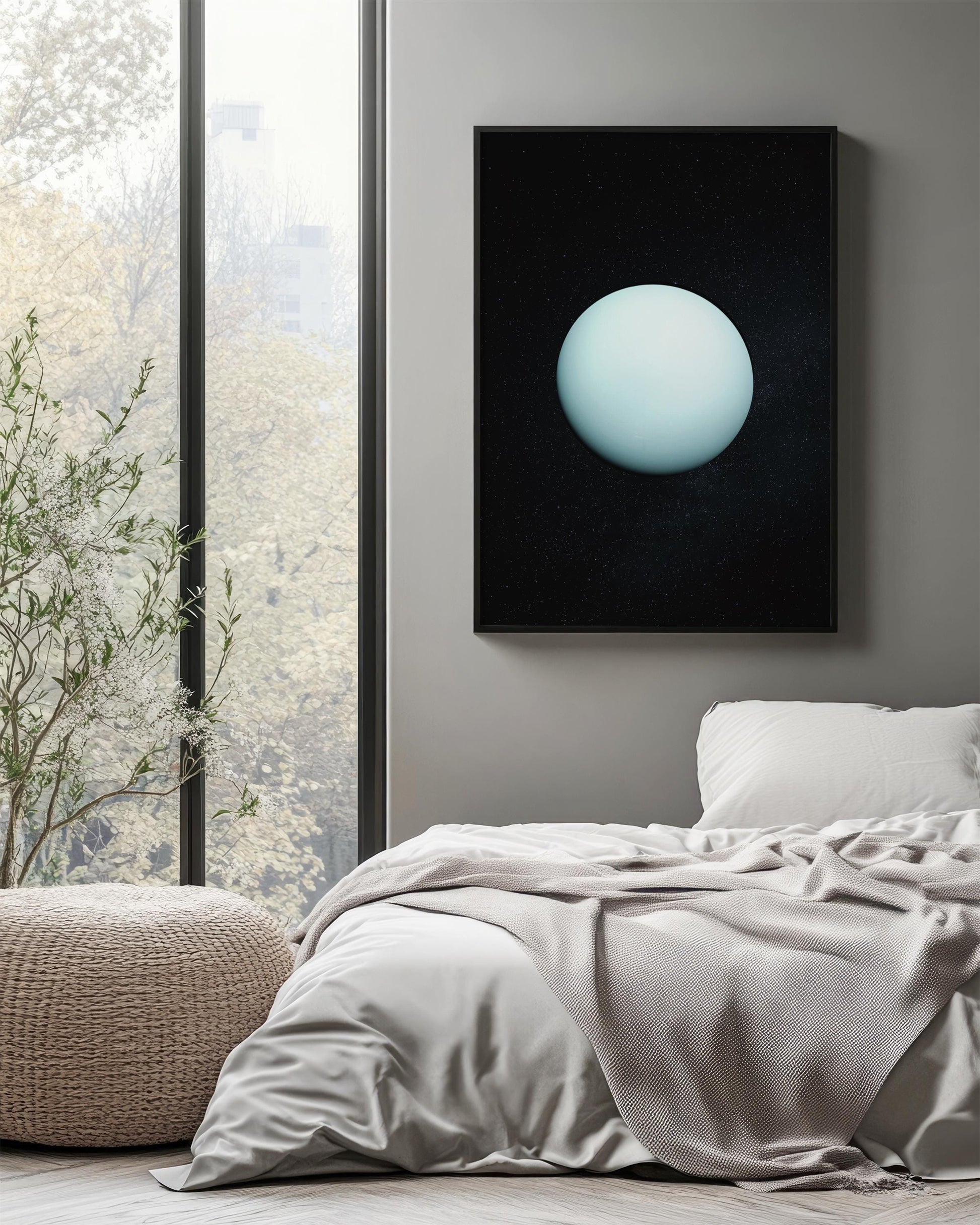 a serene bedroom scene with a large window overlooking a lush green tree outside. The window is framed by a black border, and a framed image of a blue planet is hanging on the wall above the bed. The bed is covered in a white comforter and has a single white pillow. A woven ottoman is placed next to the bed, and a potted plant is located on the floor next to the window.