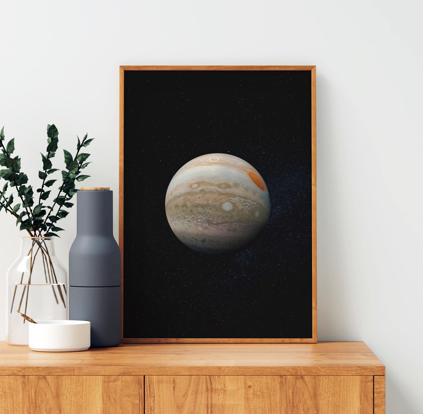 a framed picture of the planet Jupiter against a black background, placed on a wooden shelf next to a vase with green leaves and a white bowl.