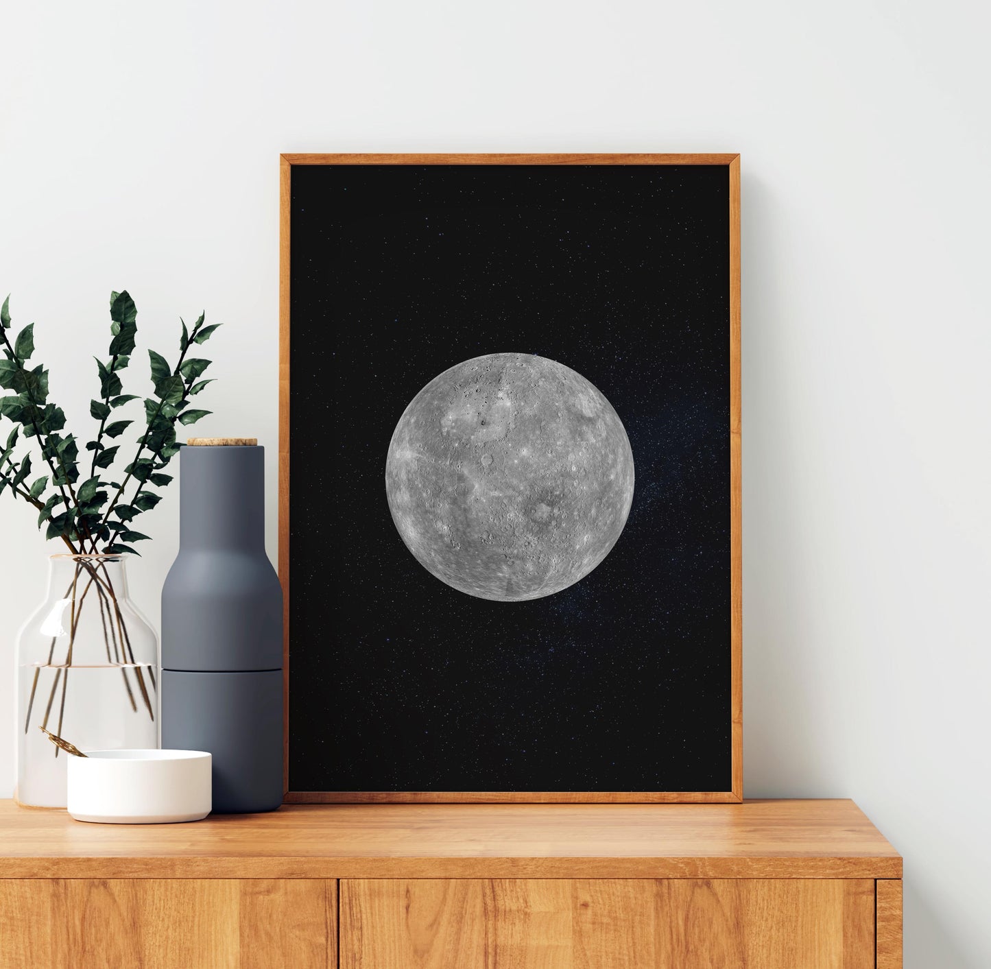 a framed picture of a moon against a black background, placed on a wooden shelf next to a vase with greenery and a white bowl.